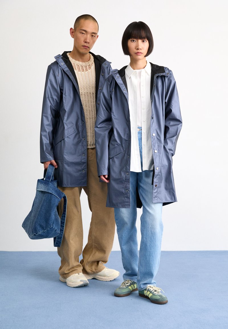 Two individuals wear matching dark blue, waterproof, hooded raincoats with snap buttons. One holds a denim tote, while the other wears a white shirt.