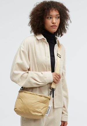 Gepolsterte beige Schultertasche aus Nylon mit glatter Textur, ausgestattet mit einem Markenetikett und verstellbarem Gurt. Kompaktes Design mit weicher Form.