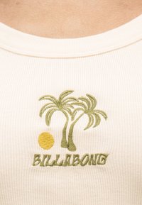 Κοντινό πλάνο σε κρεμ ριμπ ύφασμα με πράσινους κεντημένους φοίνικες, έναν κίτρινο ήλιο και το κείμενο "BILLABONG".