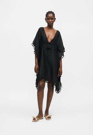 Žena nosi voľné čierne šaty s strapcami na rukávoch a leme, hlboký výstrih do V s viazaním so strapcami, stojaca v neutrálnych sandáloch na bielom pozadí.