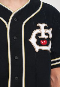 Fekete gombos ing, fehér és arany díszítéssel, amelyen egy nagyméretű hímezett logó található piros kiegészítővel. Sima textúra, baseball mez stílus.