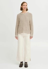 Pull en maille beige avec texture côtelée et lignes irrégulières, associé à un pantalon large blanc et des mocassins sombres avec des accents dorés.