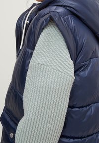 Marineblaue Steppweste mit leichten, strukturierten Strickärmeln. Verfügt über eine Kapuze und weißen Kordelzug sowie kontrastierende Materialien und Nahtdetails.