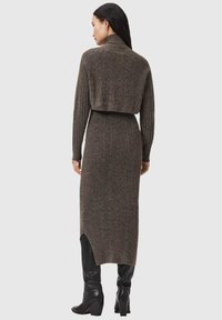 Dlhé rebrované svetrové šaty z tmavohnedého pleteného materiálu s vysokým golierom a rozparkom na leme, doplnené čiernymi čižmami po kolená.