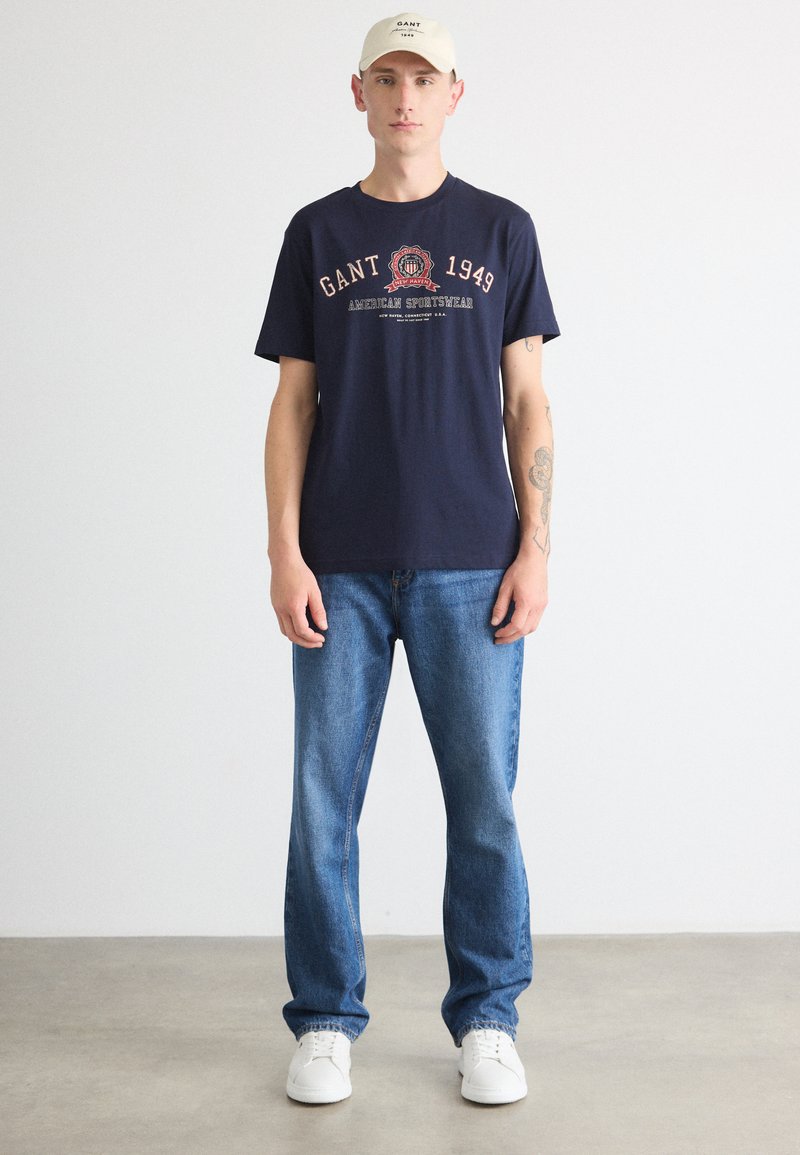 Granatowa bawełniana koszulka z grafiką tekstową, zestawiona z jasnoniebieskimi dżinsami i białymi trampkami. Dodatki to beżowa czapka z logo.