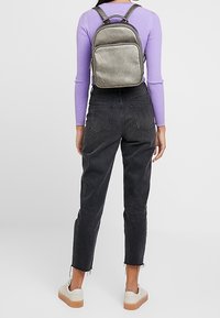 Metaliczny srebrny plecak o prostokątnym kształcie, z zamykaną na suwak kieszenią i regulowanymi paskami, noszony na fioletowej bluzce i czarnych spodniach.