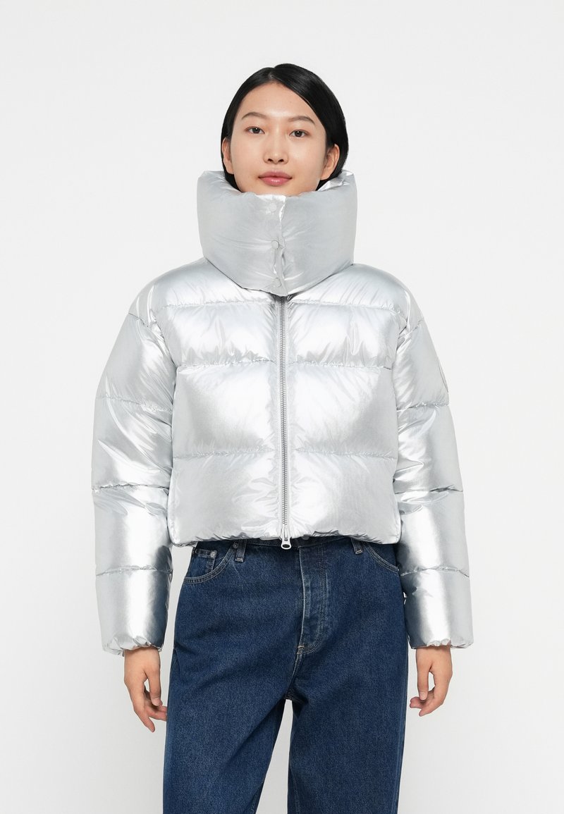 Silver puffer jacket with a high collar, shiny material, horizontal quilted pattern, and front zip closure; paired with dark denim pants.