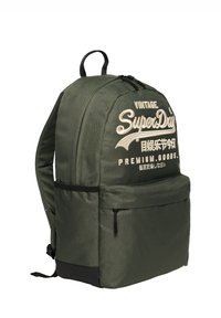 Mochila Superdry de color verde oscuro con texto bordado, bolsillo frontal con cremallera y panel base negro, vista en ángulo sobre fondo blanco.