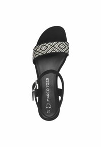 Sandalia negra con una tira trenzada texturizada que presenta un patrón de diamantes. Diseño de punta abierta y una cinta delgada en el tobillo con un hebilla de metal.