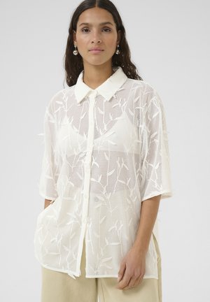 Camicia bianca a strati realizzata in tessuto a rete con motivi a foglia, colletto, maniche corte, chiusura anteriore con bottoni e tasche laterali.