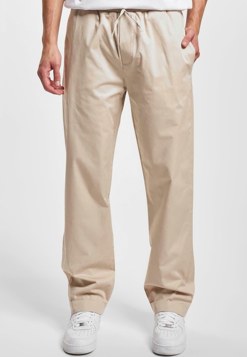 Pantaloni bej cu picioare largi, cu un talie elastică, șnur și buzunare laterale. Purtați cu adidași albi, textură de țesătură netedă.