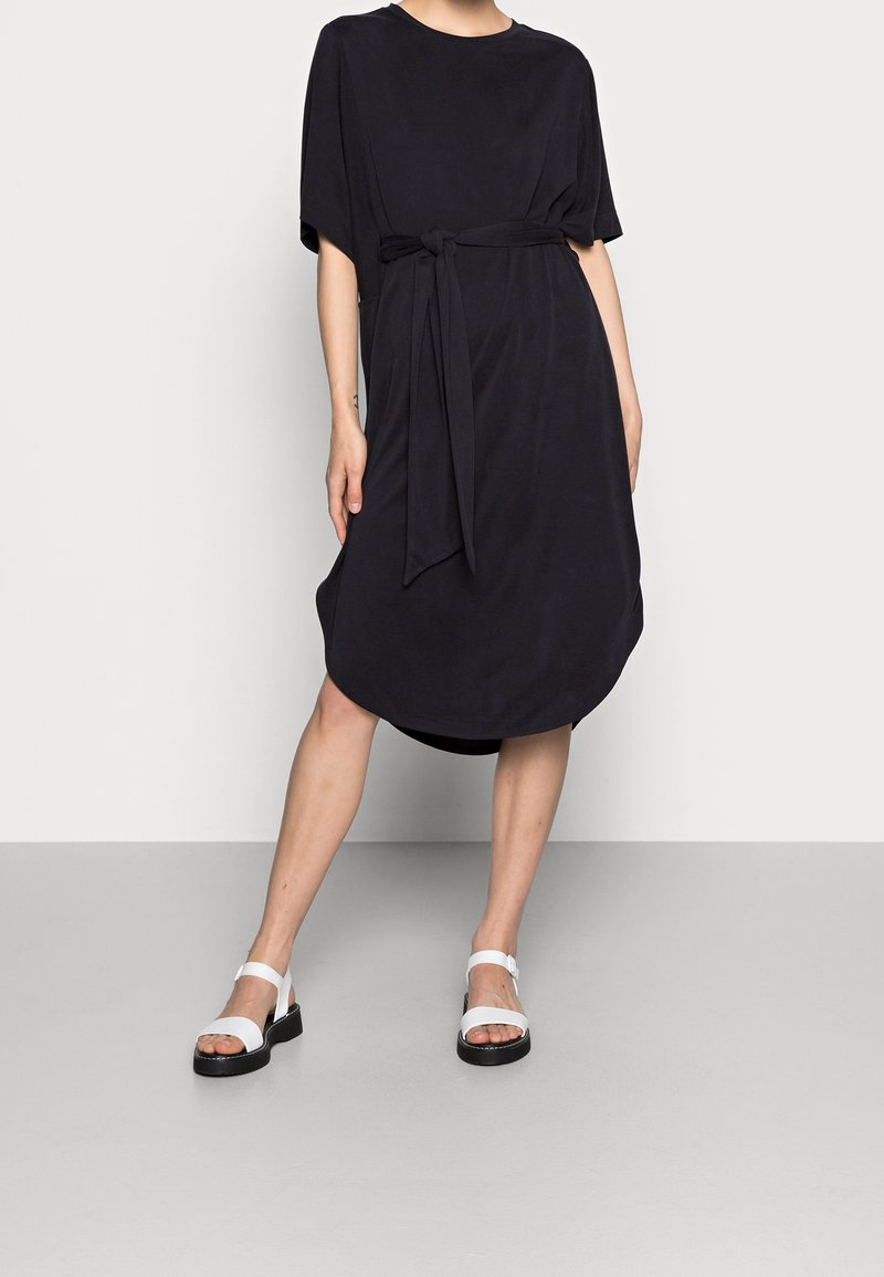 Personne portant une robe noire ample jusqu’aux genoux avec ceinture à la taille et des sandales blanches, debout les jambes écartées devant un fond uni.