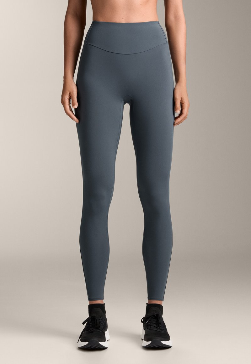Hochtaillierte, enganliegende Leggings in einem gedämpften Grauton mit glattem Texture. Sie verfügt über einen nahtlosen Bund und wird mit schwarzen Turnschuhen ergänzt.