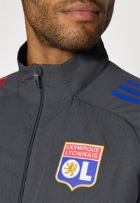 Grå treningsjakke med høy krage, med røde og blå skuldrestreker, og et brodert Olympique Lyonnais-logo på brystet.