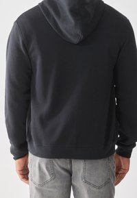 Schwarzer Kapuzenpullover aus weichem Material mit lockerer Passform, ausgestattet mit einer Kängurutasche und gerippten Bündchen. Getragen mit grauen Jeans.