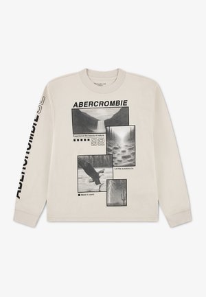 Chemise beige à manches longues avec des photos noires sur le thème de la nature, texte "ABERCROMBIE" et phrases comme "Experience the beauty of nature" sur le devant et la manche.