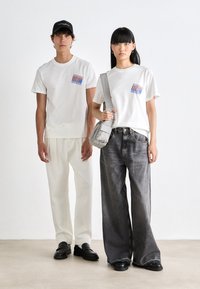 Two models wearing white graphic T-shirts; one in white trousers and black cap, the other in wide black jeans, carrying a gray shoulder bag.