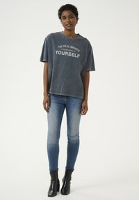 Per didelė pilka T-shirt su balta užrašu "TIKRAI AUGIMAS YRA BŪTI DAUGIAU SAVIMIS", derinama su šviesiai mėlynais siaurais džinsais ir juodais kulkšnies botais.