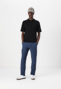 Svart polo-skjorte, blå bukser og hvite sneakers. Modellen har på seg en grå caps. Utstyret har en uformell, avslappet passform. Minimale designelementer.