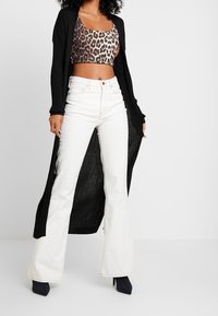 Lång svart ribbad kofta över en leopardmönstrad crop top, med vita utsvängda jeans och svarta spetsiga klackar.