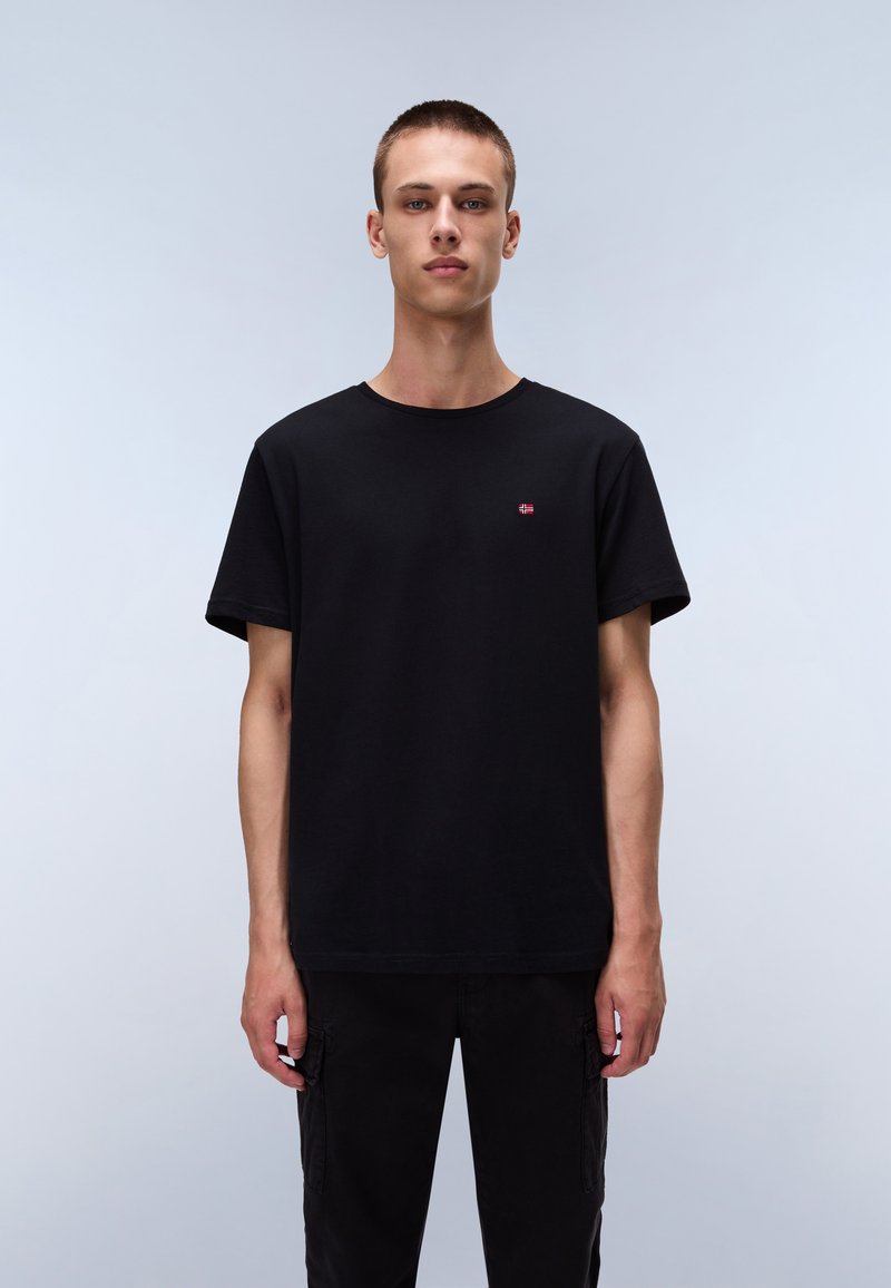 T-shirt en coton noir à manches courtes, col rond et petit logo rouge sur la poitrine gauche. Porté avec un pantalon cargo noir.