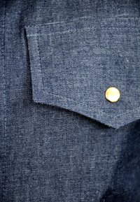 Närbild av en blå denimskjortficka med synlig sömnad och en enda rund mässingsknapp.