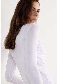 Vrouw met lang bruin haar die een nauwsluitend wit shirt met lange mouwen draagt, kijkt over haar schouder tegen een effen witte achtergrond.