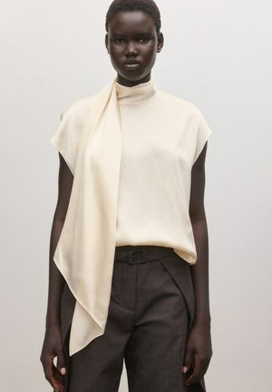 Donna con una blusa senza maniche color crema con dettaglio a sciarpa drappeggiata e pantaloni sartoriali a vita alta grigio scuro, su uno sfondo semplice.