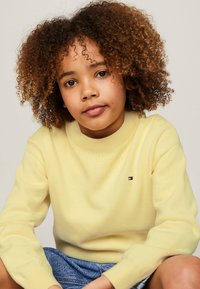Maglione giallo lavorato a maglia con collo a giro ribattuto e piccolo logo ricamato sul petto, indossato sopra pantaloni corti di jeans. Tessuto texturizzato.