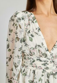 Abito floreale con scollo a V profondo, caratterizzato da uno sfondo crema decorato con foglie verdi e fiori rosa; tessuto texturizzato con maniche arricciate.
