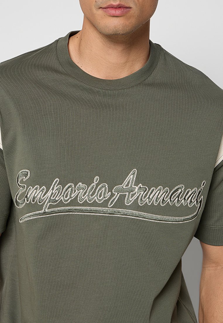 Hombre con camiseta verde oliva con "Emporio Armani" bordado en cursiva en el pecho.