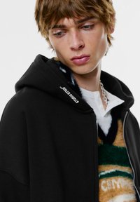 Fekete cipzáras kapucnis pulóver puha textúrával, kapucnival és logó részlettel. Zöld, bézs és fehér mintás pulóver fölé rétegezve.