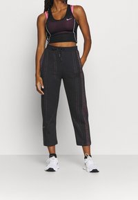 Frau in schwarzem Cropped Top mit rosa Trägern, schwarzen, kurzen Sporthosens mit roter Naht und weißen Turnschuhen, die an einer weißen Wand steht.