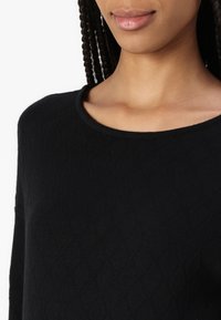 Svart genser med et teksturert diamantmønster, rund hals og lange ermer. Stoffet ser mykt og strikket ut. Nært bilde.