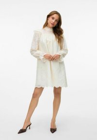 Robe mini en dentelle blanche avec des manches longues en tulle, un col à volants et un ourlet en forme de vagues. Le modèle l'associe avec des talons aiguille brun foncé.