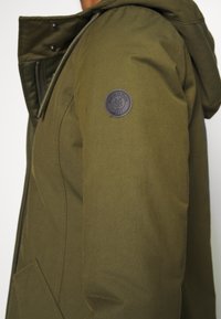 Olijfgroene jacket van een gestructureerde stof, met een rits en drukknoopsluiting, een verstelbare capuchon en een circulair zwart logo op de mouw.