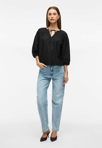 Melna, brīvi pieguļoša blūze ar kakla saiti, puffētām piedurknēm; apvienota ar augstu vidukļa viegli ziliem džinsiem un vīna sarkaniem apaviem.