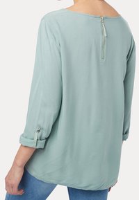 Ljusgrön blus med långa ärmar, bakdragkedja, uppvikbara ärmar fästa med en knapp, jämn textur och en avslappnad passform.