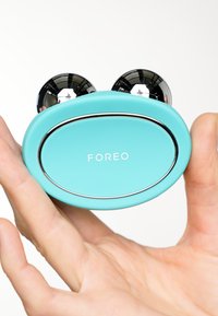 Dispositif portable turquoise avec un pourtour argenté brillant et deux sphères noires brillantes en haut. Affiche le nom de la marque "FOREO" de manière proéminente.