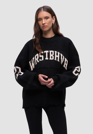 Schwarzer gestrickter Pullover in lockerer Passform, mit einem creamfarbenen Band über der Brust, auf dem der Text "WRSTBHVR" und die Nummer "23" stehen. Ausgefranster Saum.