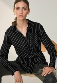 Črno-bele progaste srajce z dolgimi rokavi in žepom na prsih, ki ima ovratnik z gumbi. Model sede, oblečen v slim fit hlače.