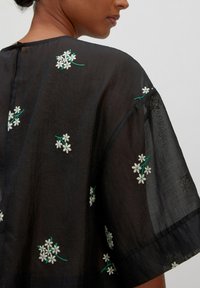 Schwarzes, durchscheinendes Kleidungsstück mit weißer und grüner floralem Stickerei. Lockerer Schnitt, weite Ärmel und eine Knopfverschluss auf der Rückseite.