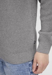 Harmaa neulottu pusero, jossa on tekstuurikuvio, ribbaatut hihat ja helma sekä pieni musta etiketti sivulla sauman lähellä.