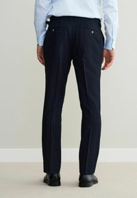 Navy pinstriped trousers made of woven fabric, featuring back pockets with metallic buttons and a tailored fit. Worn with black dress shoes.
