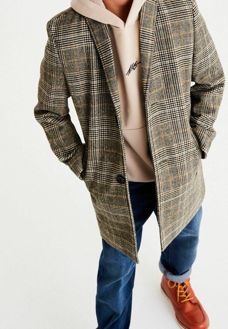 Kariertes Überhemd in Braun, Beige und Grün, mit taillierter Passform und Einzelknopfverschluss, kombiniert mit einem beigen Hoodie und blauen Jeans.