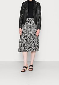 Schwarze Lederbikerjacke über einem taillierten schwarzen Oberteil, kombiniert mit einem knielangen weißen Rock mit einem schwarzen abstrakten Muster und schwarzen Absatzsandalen.