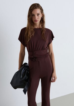 Woman with long wet hair wears a dark brown fitted jumpsuit with a flower detail at the waist, holding a large black leather bag.