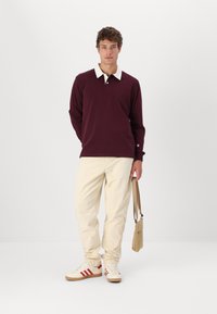 Polo shirt bordeaux a maniche lunghe con colletto bianco, abbinato a pantaloni in velluto a coste color crema. Gli accessori includono una borsa a tracolla beige e sneaker bianche.