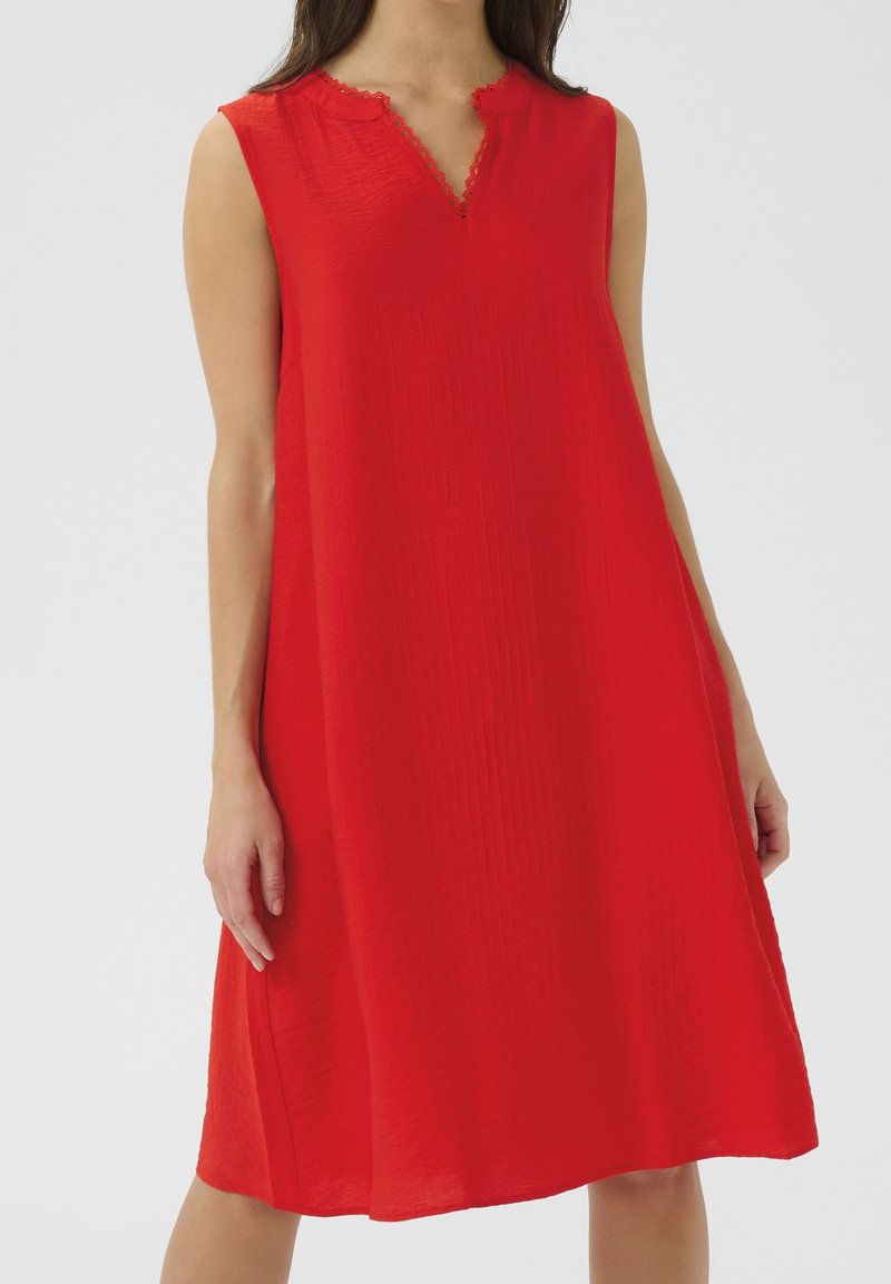 Robe rouge sans manches avec un décolleté en V orné de délicates bordures en dentelle, tombant librement juste en dessous des genoux d'une femme debout.