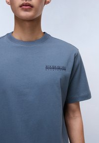Blue cotton t-shirt with short sleeves, round neckline, and a small, dark printed logo on the chest. Smooth texture, casual design.